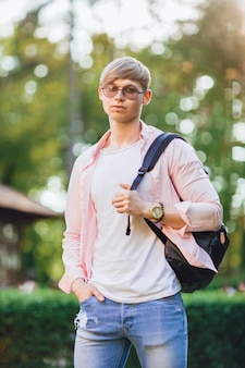 O jovem bonito em roupas casuais com óculos escuros e uma mochila está no campus