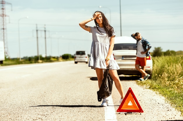 Foto grátis o jovem casal quebrou o carro durante uma viagem para descansar. eles estão tentando parar outros motoristas e pedir ajuda ou pedir carona. relacionamento, problemas na estrada, férias.