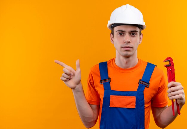 O jovem construtor usando uniforme de construção e capacete de segurança segura um alicate de ranhura e aponta para a esquerda com o polegar