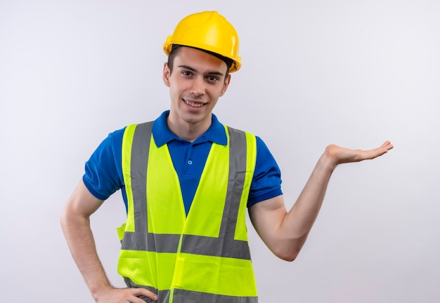 O jovem construtor usando uniforme de construção e capacete de segurança sorri e aponta para a esquerda com a mão