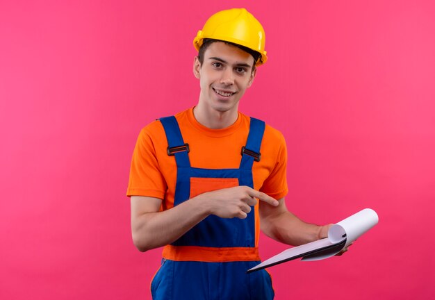 O jovem construtor vestindo uniforme de construção e capacete de segurança sorri e aponta com o polegar na prancheta