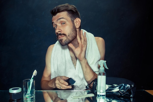 O jovem no quarto sentado em frente ao espelho coçando a barba em casa. emoções humanas e conceito de estilo de vida