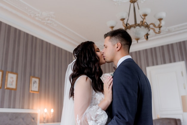 O noivo e a noiva caucasiana estão beijando ternamente no quarto de hotel claro