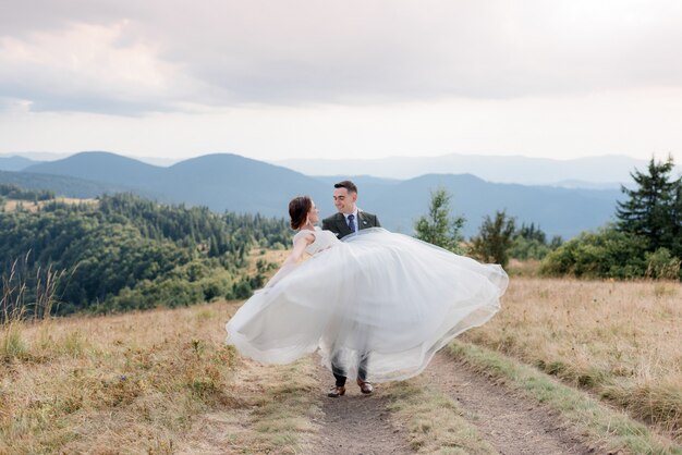 O noivo sorrido está carregando a noiva vestida de vestido de noiva branco no dia ensolarado de verão nas montanhas