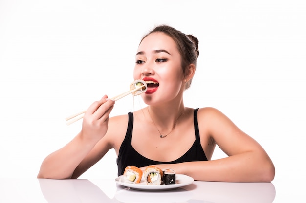 O olhar consideravelmente asiático com penteado modesto senta-se na tabela come rolos de sushi sorrindo isolado no branco