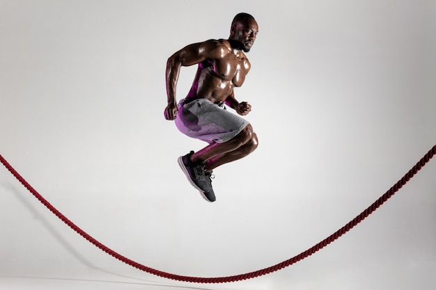 O primeiro. jovem fisiculturista afro-americano treinando no fundo cinza do estúdio. modelo masculino único musculoso em roupas esportivas, saltando sobre a corda de batalha. conceito de esporte, musculação, estilo de vida saudável.