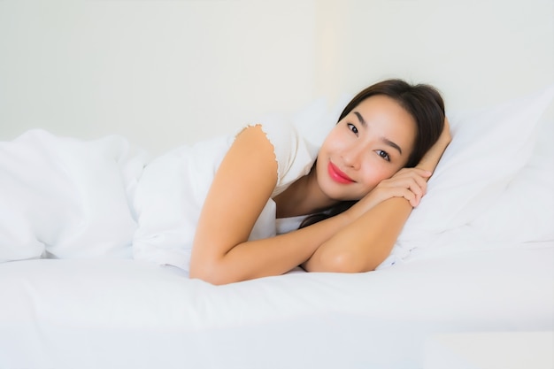 O retrato a mulher asiática nova bonita relaxa o sorriso feliz na cama com o cobertor branco do travesseiro