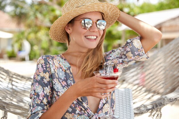 O retrato ao ar livre de uma linda mulher se sente relaxado enquanto bebe um coquetel fresco, usa chapéu e óculos de sol da moda, senta-se perto da rede e tem um bom descanso durante o tempo ensolarado de verão. Conceito de recreação