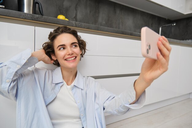 O retrato da jovem senta-se no chão da cozinha com o telefone e tira selfie no smartphone com app fil