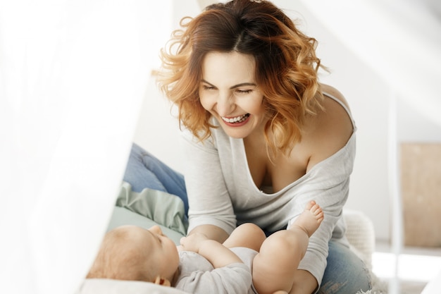 O retrato da mãe atrativa nova ri e jogando com criança recém-nascida no quarto claro confortável. manhãs quentes com a família. infância feliz.