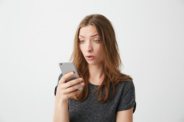 O retrato da menina bonita nova surpreendida que olha a tela do telefone com a boca aberta que surfa wed o Internet.