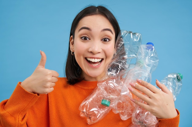 Foto grátis o retrato da menina coreana alegre mostra os polegares para cima e as garrafas plásticas recomendam a reciclagem na triagem