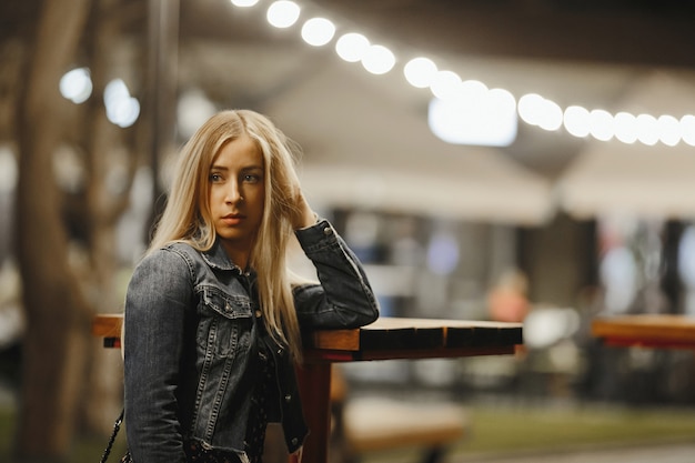 O retrato de uma menina caucasiano loura nova atrativa perto da mesa de centro alta exterior está olhando seriamente ao lado vestido no revestimento da calças de brim sob a iluminação do festão da noite