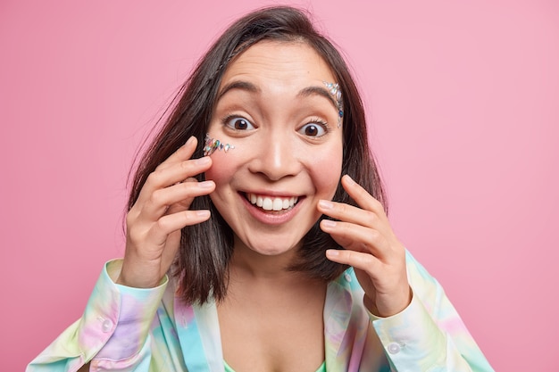 O retrato de uma mulher asiática alegre com um sorriso cheio de dentes gosta de ouvir notícias positivas, toca na pele fresca, tem pedras brilhantes coladas no rosto, expressa emoções autênticas e felizes isoladas sobre uma parede rosa