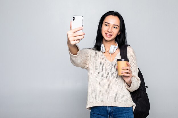 O retrato de uma mulher atrativa de sorriso que toma um selfie ao guardar retira a xícara de café isolada sobre a parede branca