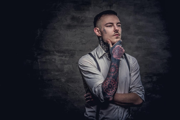 O retrato do close-up de um jovem tatuado antiquado pensativo vestindo camisa branca e suspensórios segura a mão no queixo. Isolado em um fundo escuro.