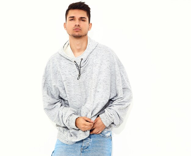 O retrato do homem modelo considerável novo vestiu-se na roupa ocasional cinzenta do hoodie que levanta na parede branca. Isolado