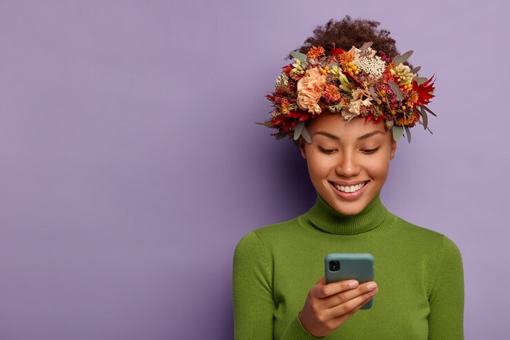 Foto grátis o retrato do modelo de outono feliz usa uma coroa de flores outonal decorativa, focado no dispositivo smartphone, lê boas notícias online, tem uma expressão de rosto feliz, modelos sobre a parede roxa do estúdio.