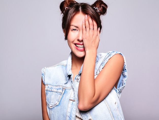 O retrato do modelo moreno bonito bonito da mulher na roupa ocasional das calças de brim do verão sem a composição com penteado dos chifres isolado no cinza. cobrindo o rosto com a mão