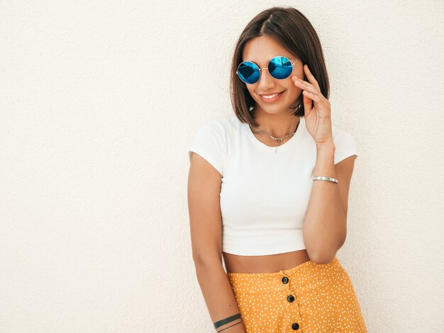 O retrato do modelo moreno sorridente bonito vestiu-se com roupas de hipster de verão. Menina na moda posando na rua perto da parede branca. Mulher engraçada e positiva se divertindo