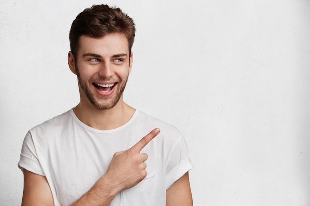 O retrato horizontal de um homem feliz de olhos azuis com a barba por fazer em uma camiseta branca casual indica com o dedo indicador no espaço em branco da cópia, anuncia algo, tem uma expressão positiva. Pessoas, conceito de propaganda