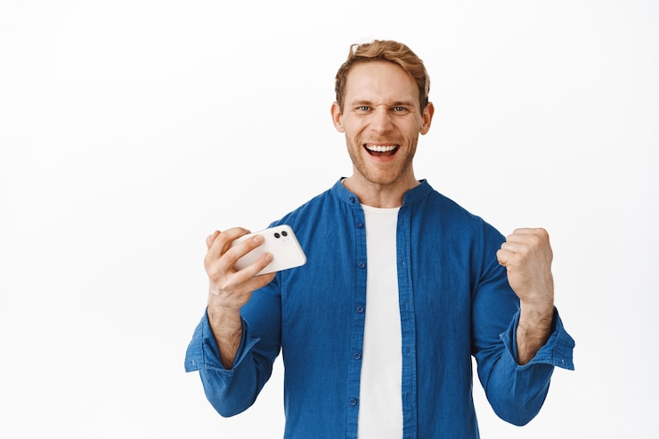Foto grátis o ruivo feliz diz sim, cerrar os punhos ao vencer no celular, triunfar, atingir a meta de sucesso no videogame para smartphone, em pé contra uma parede branca