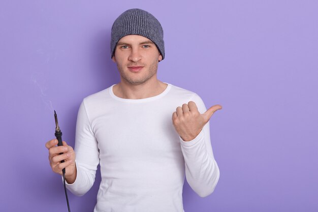 O técnico novo que está pronto para soldar o fio, o homem atrativo veste a camisa ocasional branca e o tampão cinzento guarda o ferro de solda em uma mão e aponte-o com outro polegar, isolado no roxo.