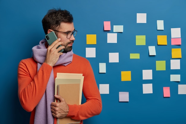 O trabalhador criativo com barba liga para alguém pelo celular, usa óculos, suéter com lenço, olha de lado nas notas coloridas coladas na parede azul