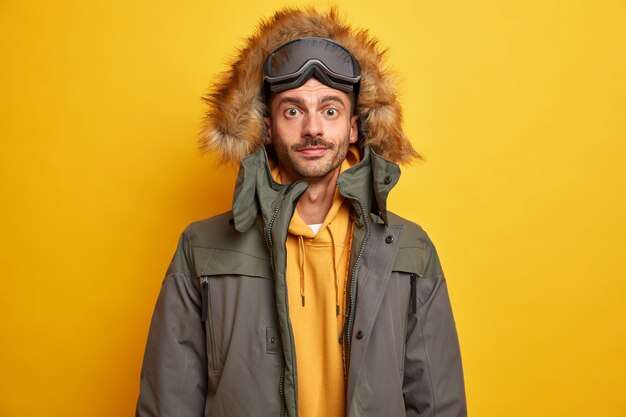 O turista sério de homem com barba por fazer descansa durante o inverno nas montanhas e gosta de praticar snowboard vestido com uma jaqueta quente com capuz de pele parece com confiança.