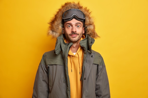 O turista sério de homem com barba por fazer descansa durante o inverno nas montanhas e gosta de praticar snowboard vestido com uma jaqueta quente com capuz de pele parece com confiança.