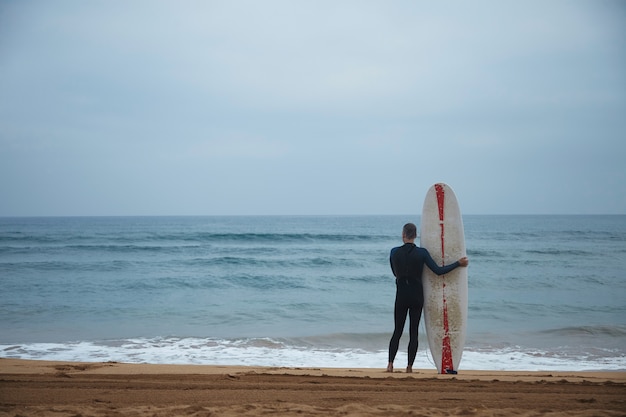 O velho surfista com seu longboard fica sozinho na praia em frente ao oceano e observa as ondas no oceano antes de ir surfar, vestindo roupa de neoprene completa no início da manhã