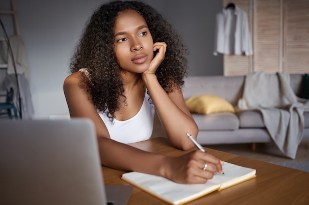 Ocupação, trabalho, tecnologia e conexão. mulher jovem e pensativa de pele escura, pensativa e com cabelos volumosos, usando o laptop para trabalho remoto, escrevendo no caderno, tentando se lembrar de algo