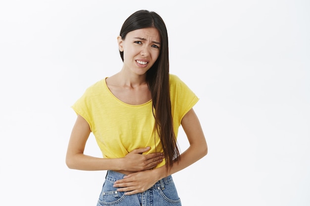 Foto grátis odeio cólicas menstruais. retrato de uma jovem fofa triste e descontente em uma camiseta amarela sentindo desconforto no estômago