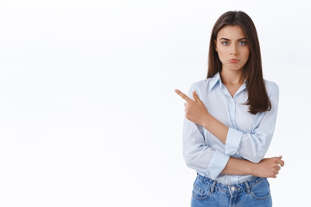 Ofendida, amuada, linda garota caucasiana de blusa azul, apontando o dedo para a esquerda, fazendo beicinho e olhando para a câmera com lamentação insultada ou expressão de reclamação em pé desapontado e descontente