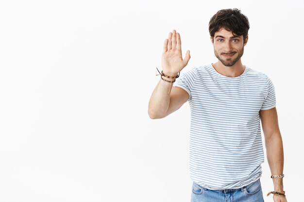 Oi amigo dê mais cinco. Retrato de um homem jovem, simpático e simpático, com olhos azuis e eriçados, levantando o braço para ondular e dizer olá, sorrindo gentilmente na frente sobre a parede branca