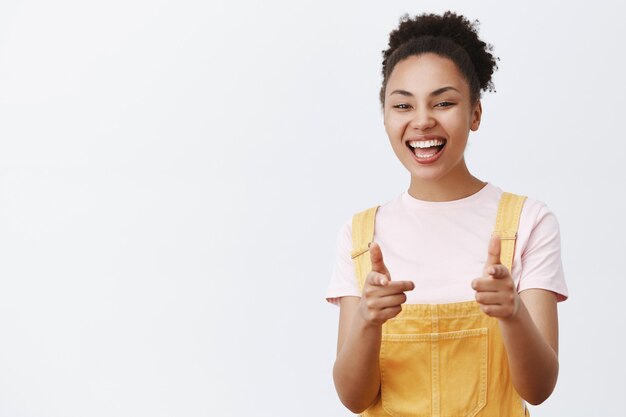 Oi te conheço. Retrato de uma adolescente afro-americana amigável, otimista e gentil, com um macacão amarelo da moda, apontando com o dedo um gesto e sorrindo amplamente