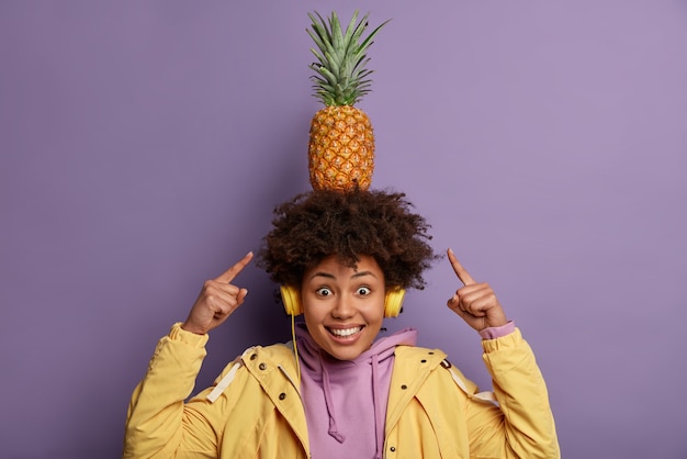 Olha como eu consigo! mulher afro-americana alegre e positiva aponta para um abacaxi na cabeça, ouve áudio em fones de ouvido, usa capuz e jaqueta, isolada sobre a parede roxa, se diverte sozinha