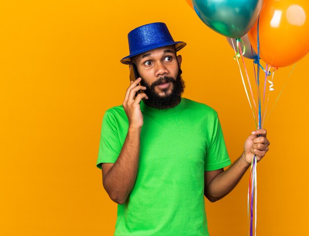 Olhando de lado, jovem afro-americano com chapéu de festa segurando balões falando no telefone isolado na parede laranja