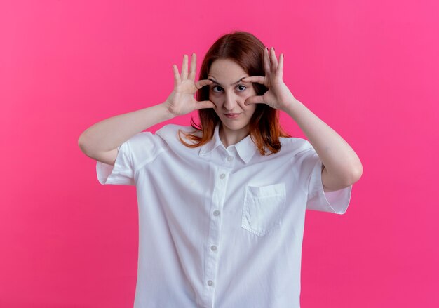 Olhando para a câmera, a jovem ruiva abrindo os olhos com as mãos isoladas em um fundo rosa