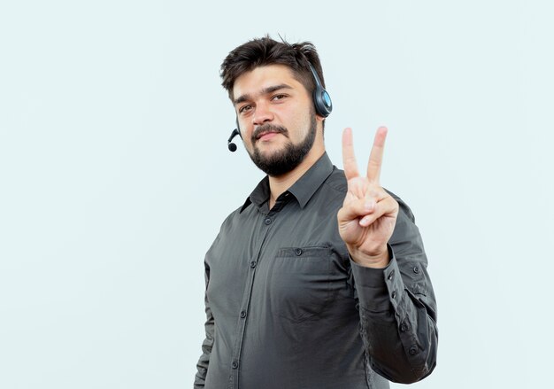 Olhando para a câmera, jovem call center, usando fone de ouvido, mostrando um gesto de paz isolado no fundo branco