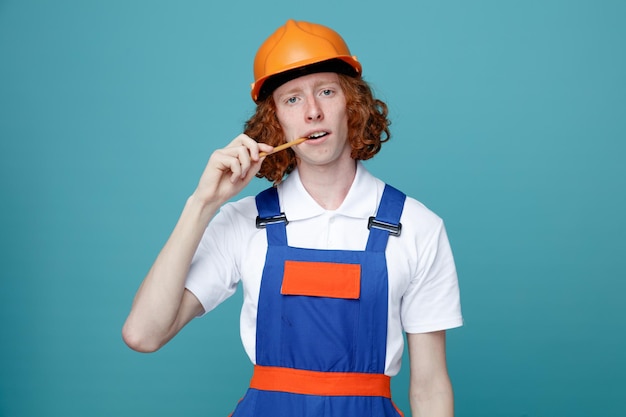 Olhando para a câmera jovem construtor de uniforme segurando o lápis na boca isolado no fundo azul