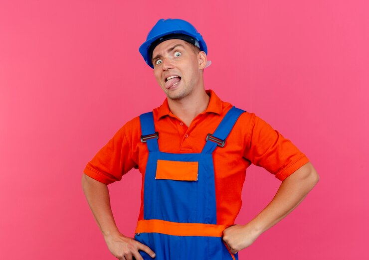 Foto grátis olhando para a câmera, jovem construtor masculino vestindo uniforme e capacete de segurança, mostrando a língua e colocando as mãos no quadril isolado no fundo rosa