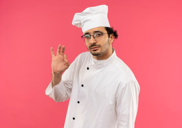 olhando para a câmera, jovem cozinheiro masculino usando uniforme de chef e óculos, mostrando um gesto de ok