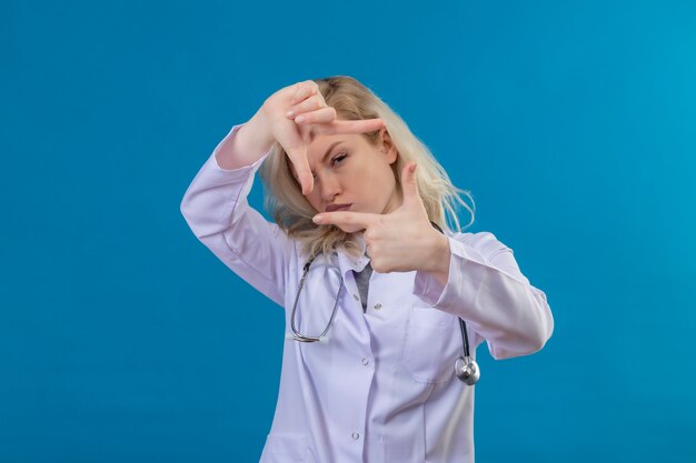Olhando para a câmera, jovem médico usando estetoscópio e vestido de médico, mostrando gesto de foto na parede azul