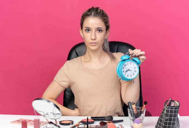 Olhando para a câmera, linda jovem sentada à mesa com ferramentas de maquiagem segurando um despertador isolado na parede rosa