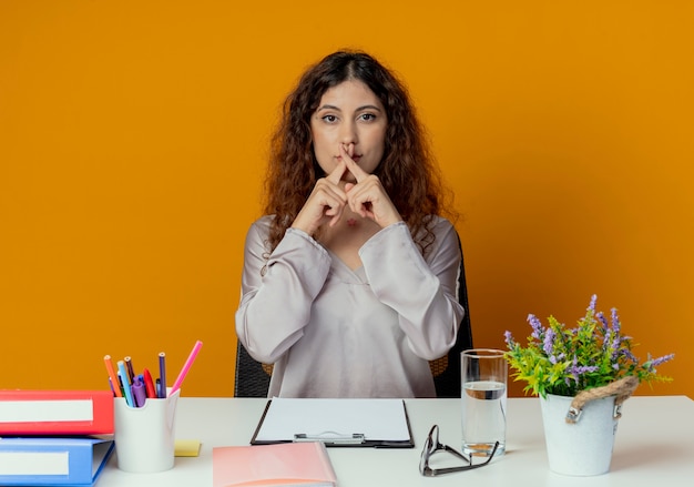 Olhando para a câmera, uma jovem bonita trabalhadora de escritório sentada à mesa com ferramentas de escritório, mostrando um gesto de não isolado em um fundo laranja