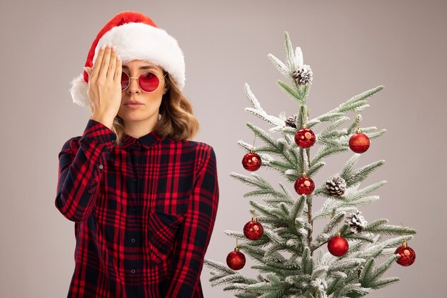 Olhando para a câmera, uma jovem linda em pé perto da árvore de Natal usando um chapéu de Natal com óculos cobertos e olhos com a mão isolada no fundo branco