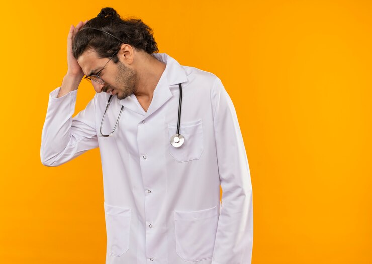 Foto grátis olhando para baixo, o jovem médico com óculos de médico vestindo manto médico com estetoscópio preocupado, colocando a mão na cabeça