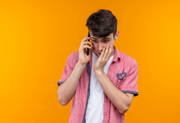 Olhando para baixo, um jovem caucasiano vestindo uma camisa rosa falando no telefone colocou a mão na bochecha na parede laranja