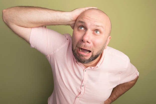 Olhando para cima com medo de um homem careca de meia-idade vestindo uma camiseta rosa e colocando a mão na cabeça isolada em verde oliva